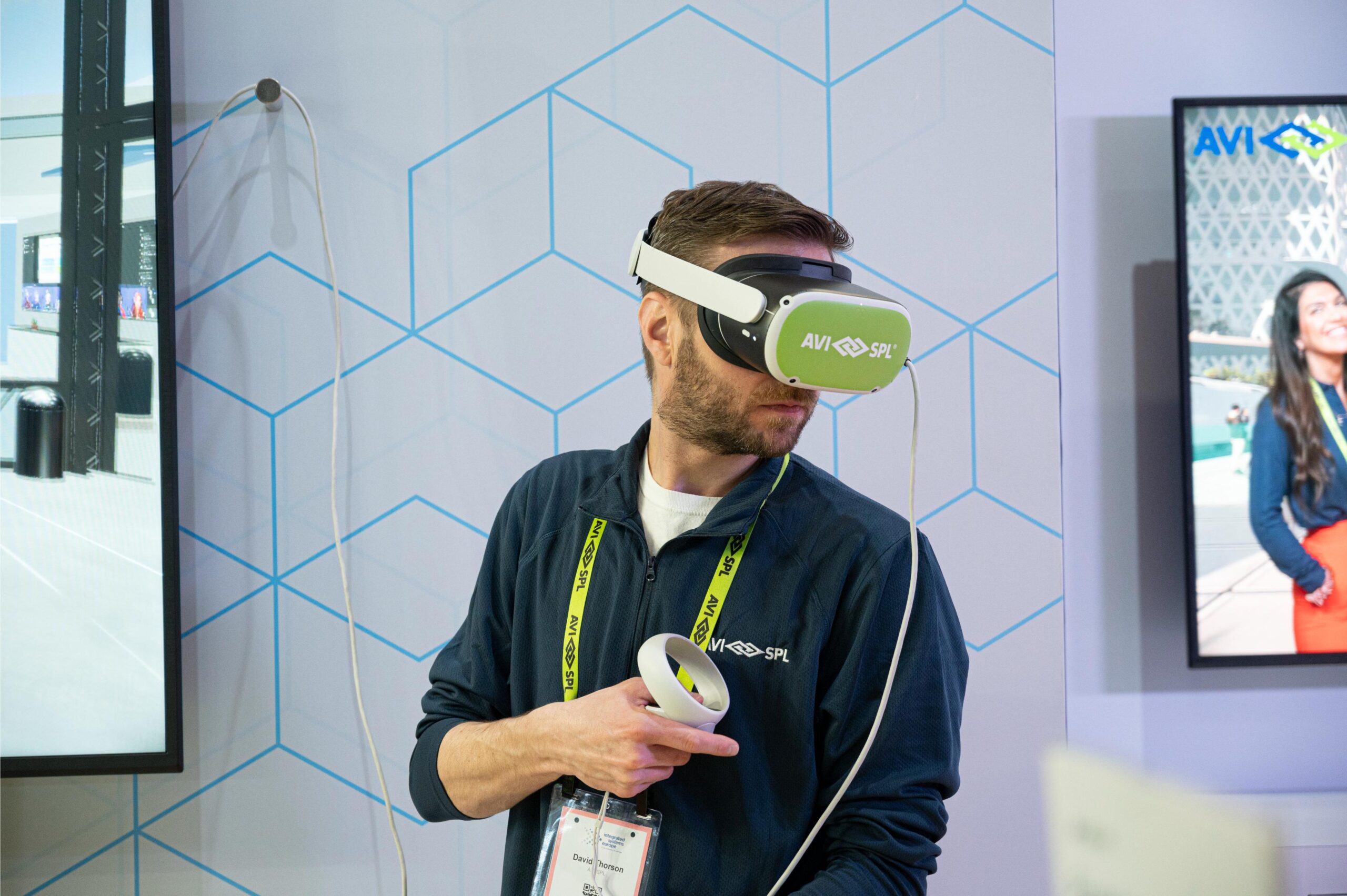 A person with a VR headset and controller, badge around neck, in front of screens displaying digital content; geometric background.