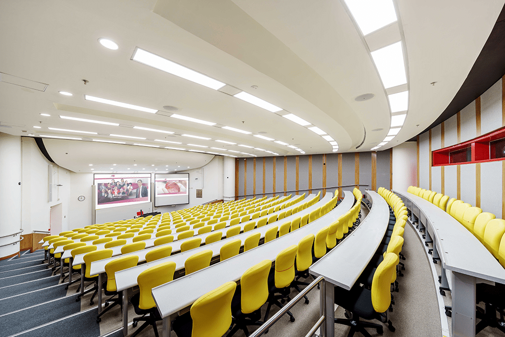 Modern lecture hall with yellow chairs, white tables, tiered seating, curved design, large screen, well-lit and airy ambiance.