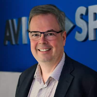 Smiling man in glasses and suit stands before blue wall with partial text AVI SP.