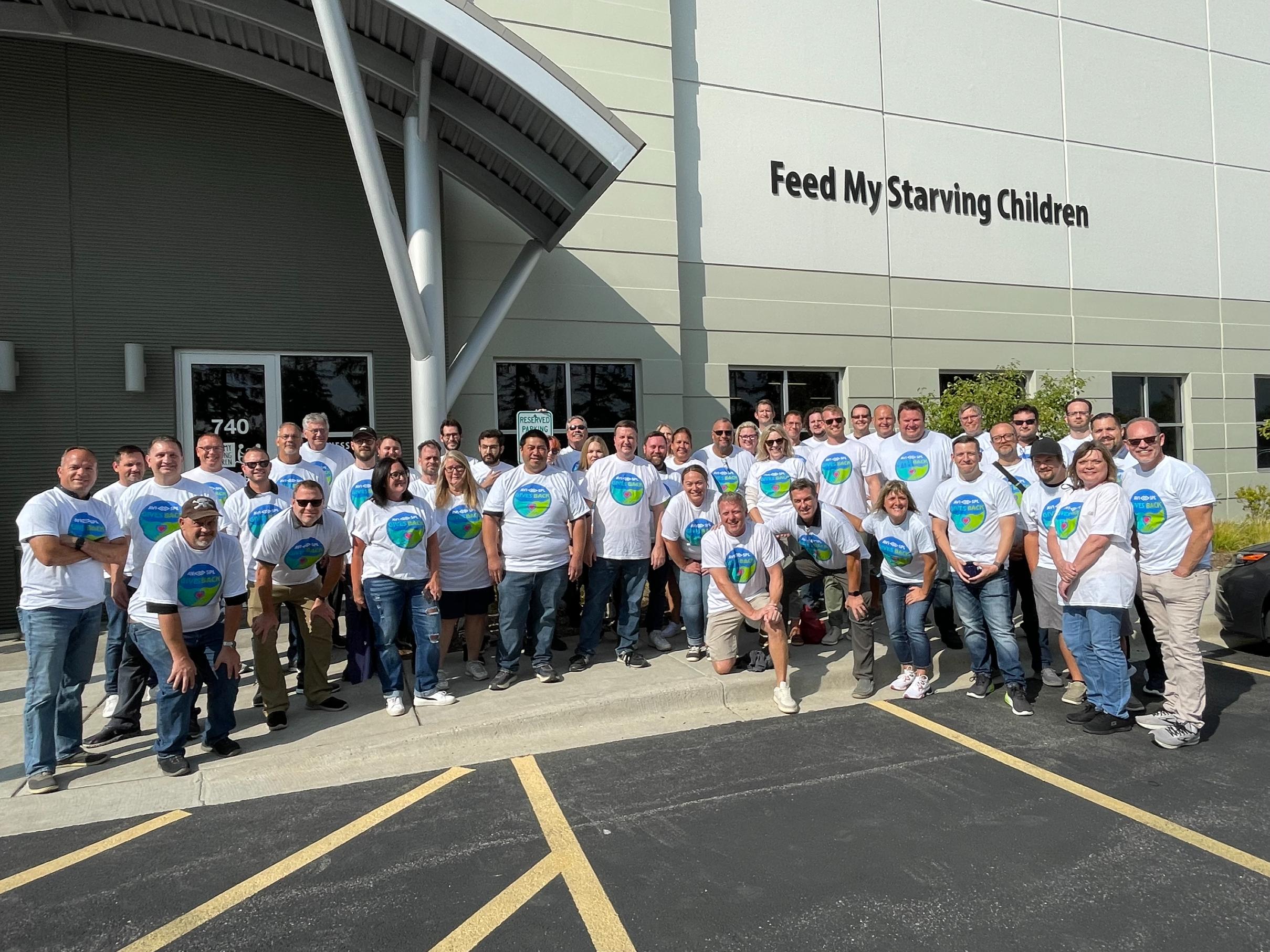 Group in matching shirts with colorful logos poses outside Feed My Starving Children, smiling under a sunny sky.