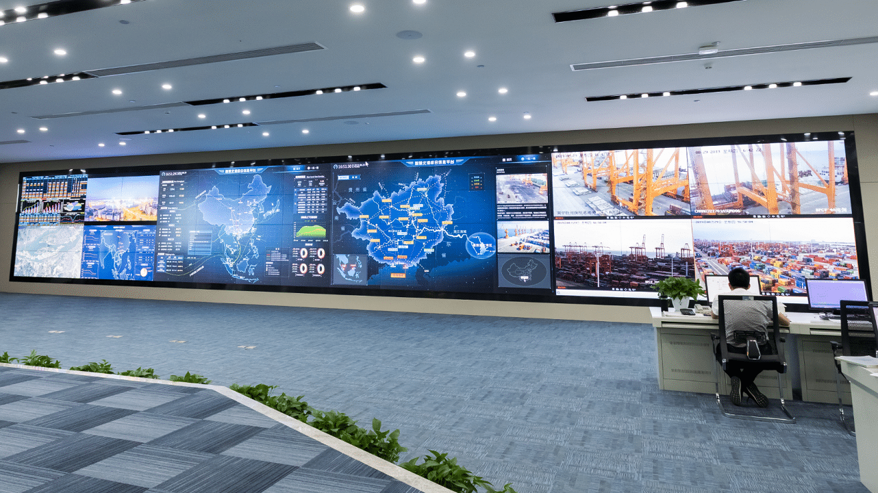 Control room with a digital screen, maps, graphs, live footage; person watching. Well-lit modern decor.