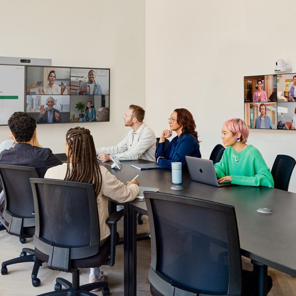 People sit around a conference table with laptops, engaged in a video call on two screens. The room is well-lit and professional.