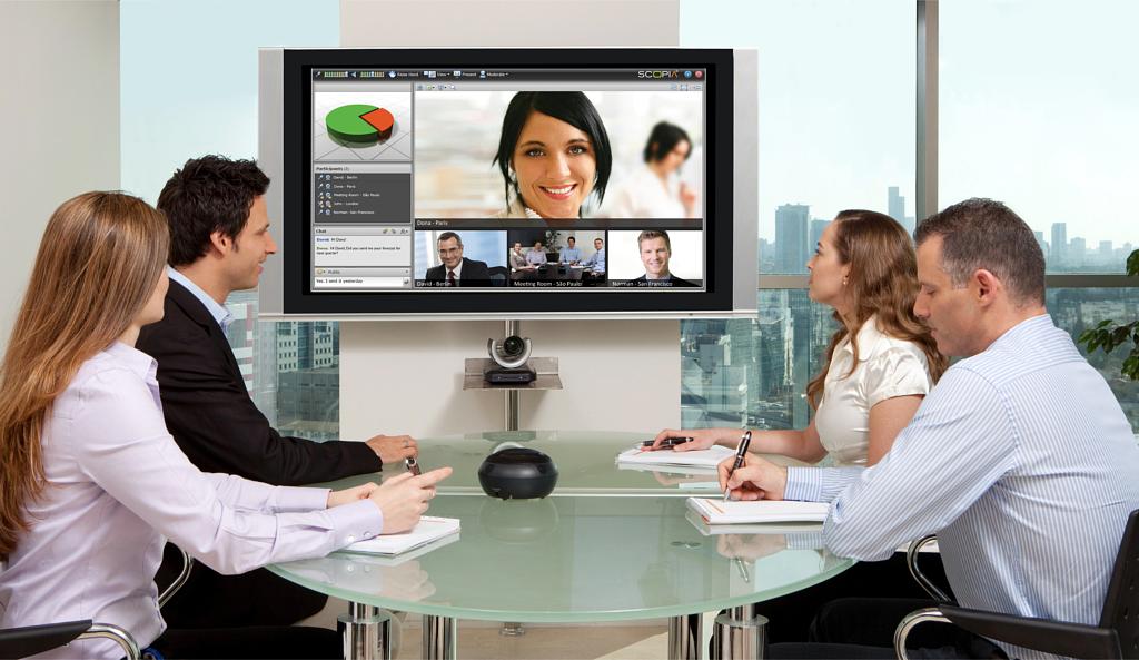 Four people at a glass table in an office video call, large screen with smiling woman; city view through big windows.