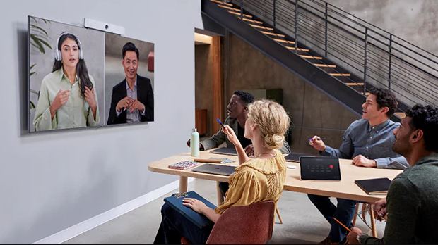 Four people sit at a modern office table for a video call; on-screen, a man and woman converse. A staircase is in the background.