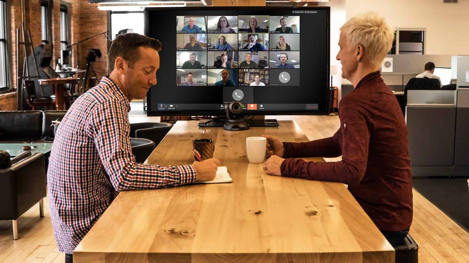 Two people at a wooden table in an office, holding mugs, focused on a video conference with multiple participants on a big screen.