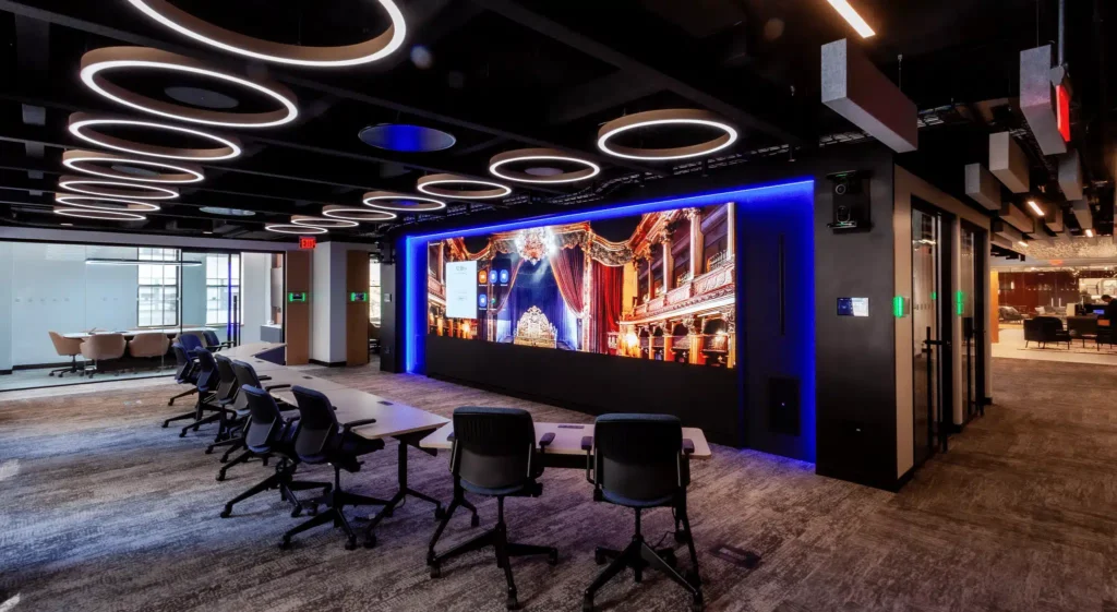 A modern conference room with a digital screen showing a theater interior, black chairs, rectangular tables, and ample lighting.