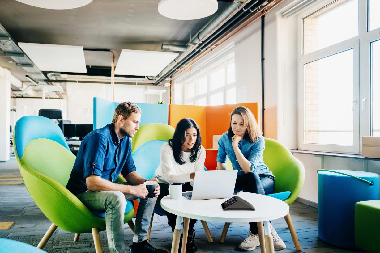 Three people discuss around a laptop at a small table in a bright modern office, seated on colorful curved chairs by a large window.