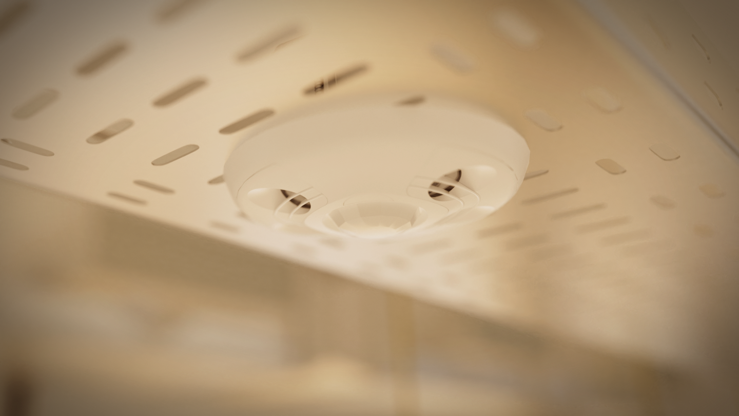 Close-up of a white smoke detector on a perforated ceiling; vents and central sensor in focus, background softly blurred.