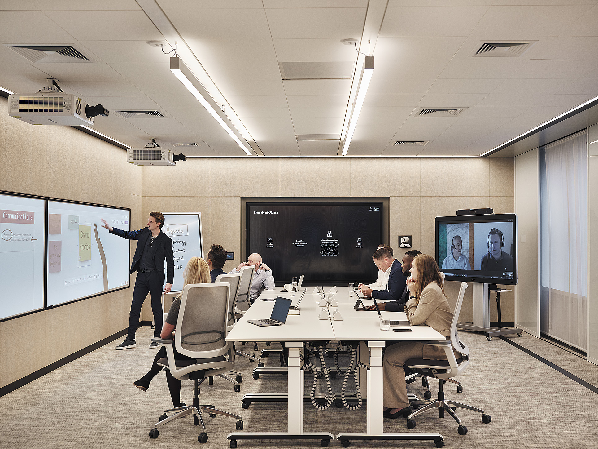 Modern conference room: people at a long table, presenter at screen, two on video call; laptops and notebooks present.