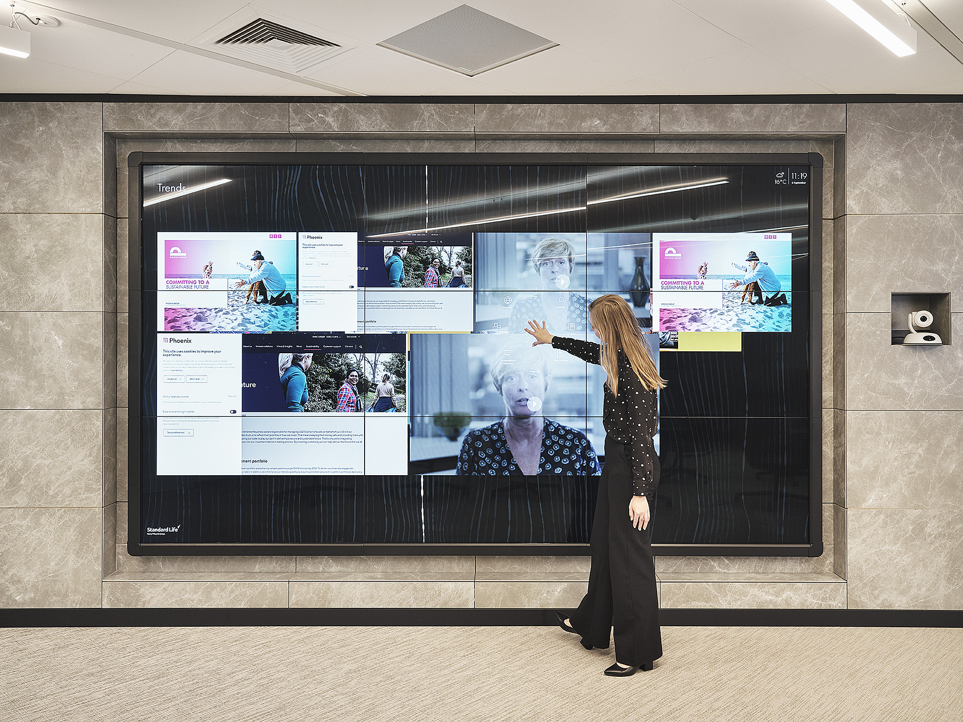 A person uses a touchscreen wall display; the modern room has gray walls, beige carpet, and a camera to the screens right.