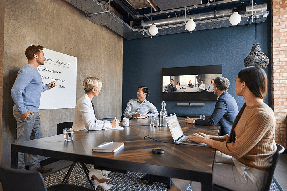 Five people in a video conference: one by the whiteboard, others seated with laptops; virtual participants on a wall screen.