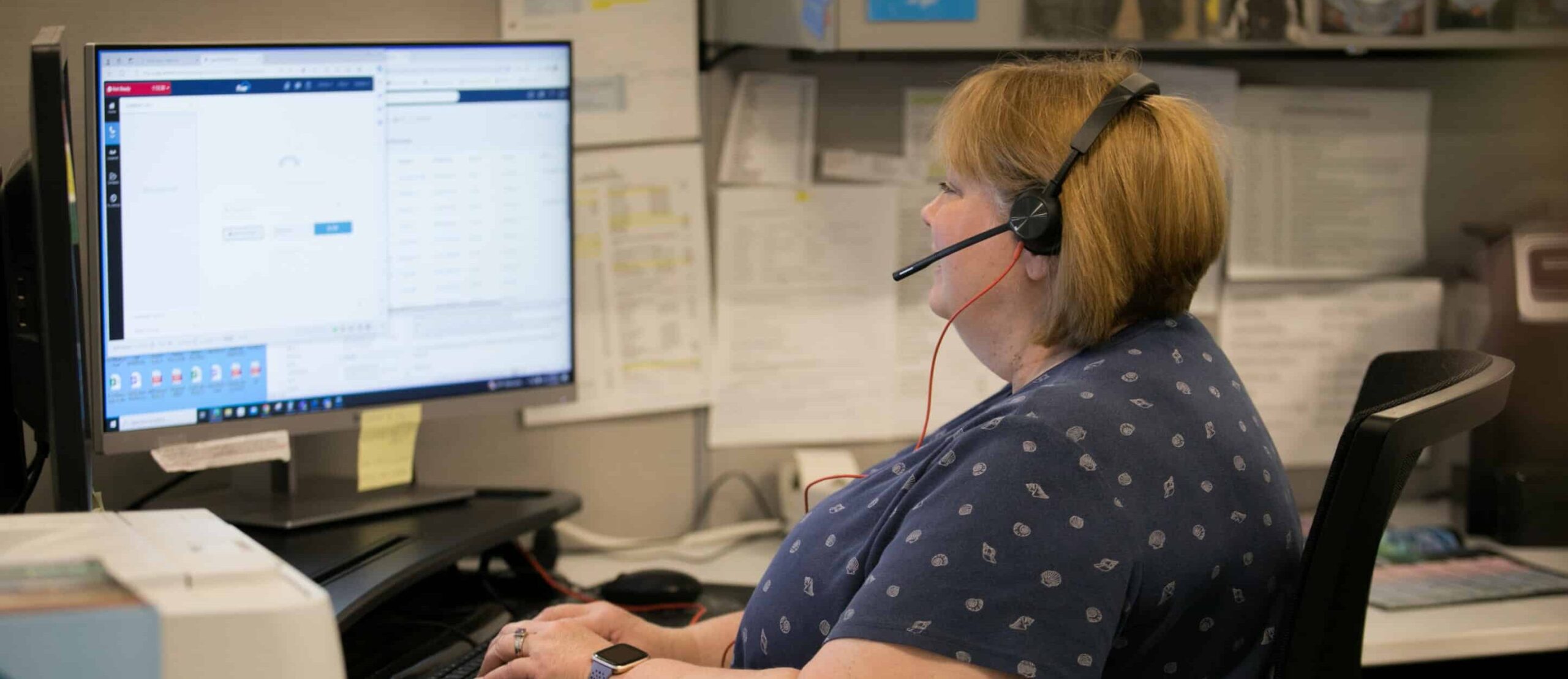 Woman wearing headset at desk with dual monitors, working on screens. Papers and office supplies visible. She wears a patterned navy shirt.