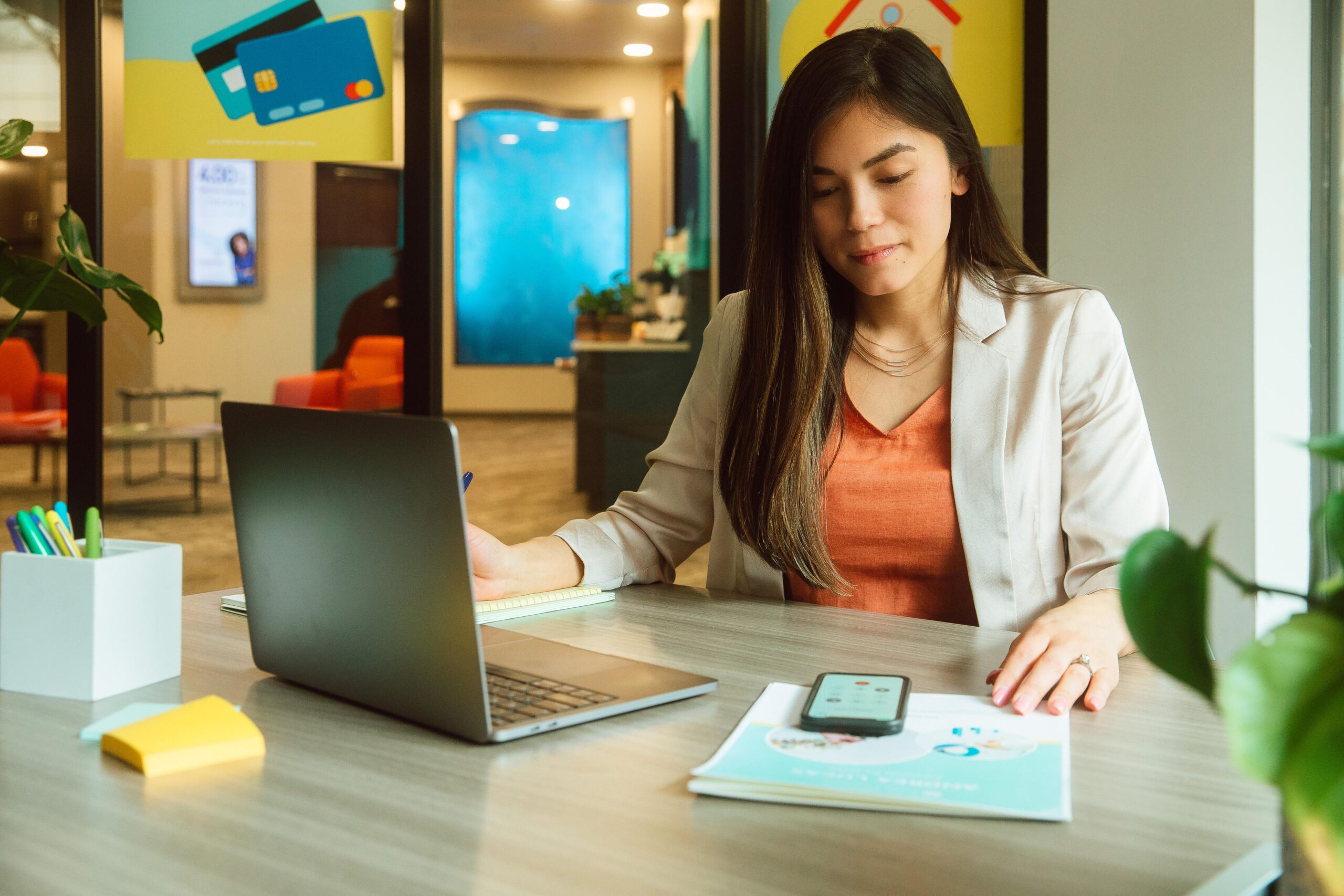 A woman at a desk uses a laptop, surrounded by documents and office supplies, focused with colorful decor in the background.