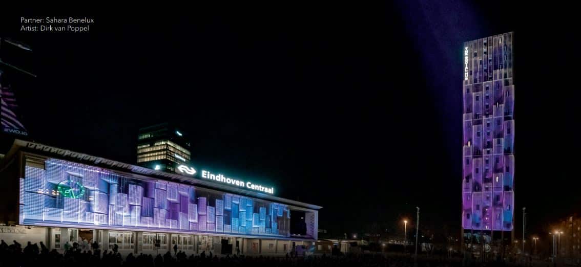 Night scene: A building with a digital art display beams blue and purple geometric patterns. Nearby, a tall illuminated tower complements the theme. Text: Eindhoven Centraal.