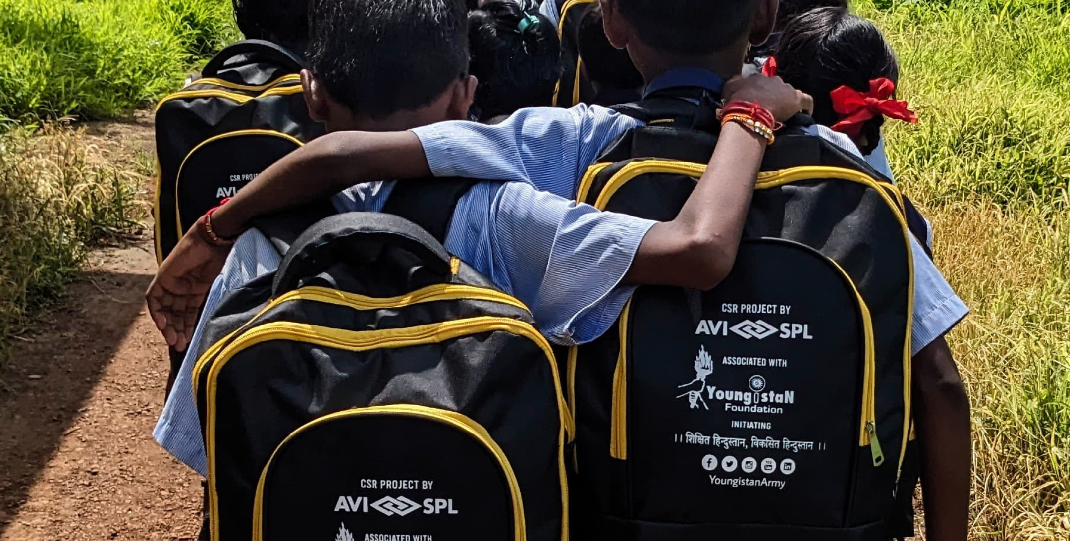 Children in school uniforms walk on a dirt path, two with arms around each other; backpacks display logos, with green grass behind.