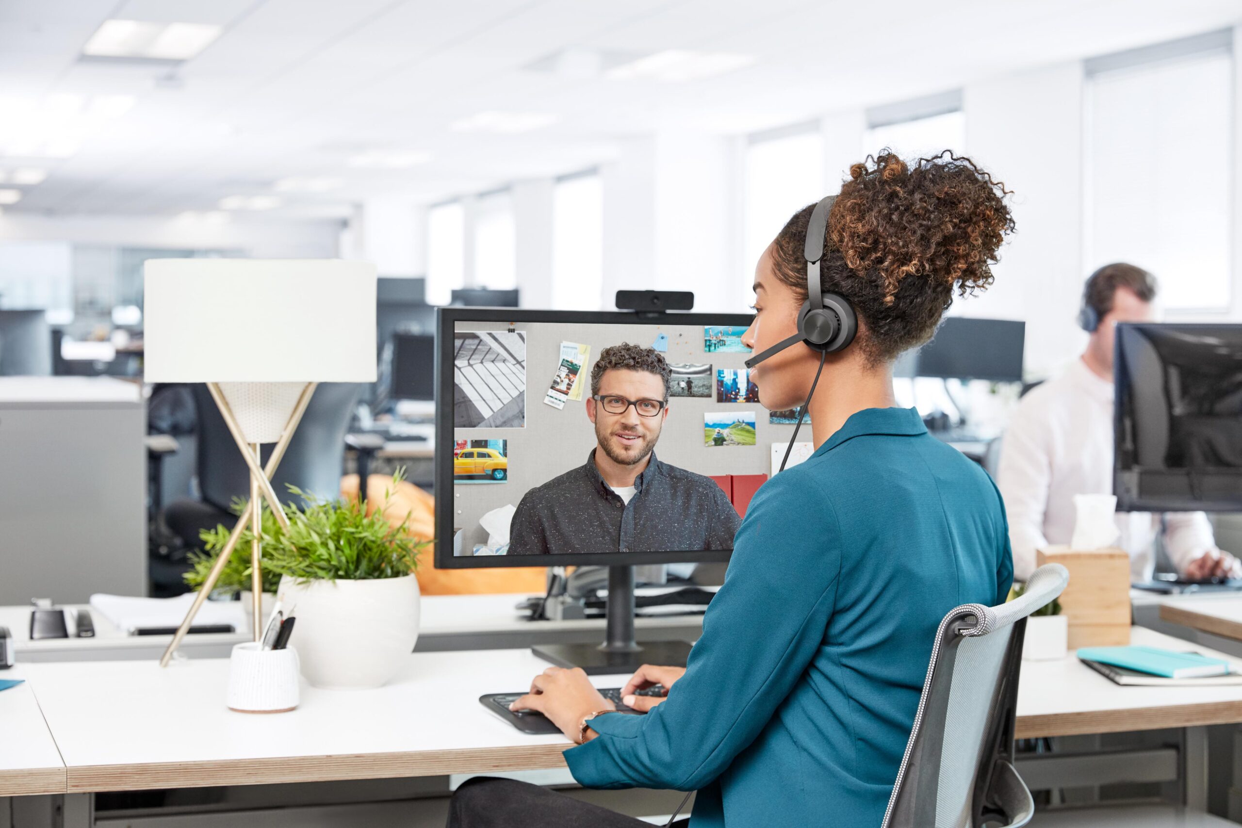 woman in blue shirt with headset on video call, man visible on screen; bright, organized office.
