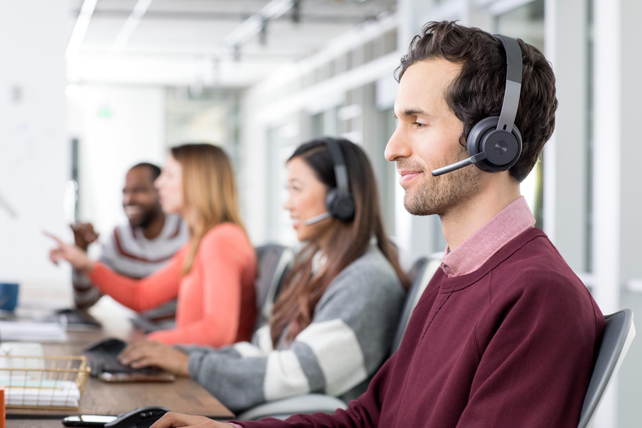 People sit at desks in a bright office, wearing headsets. A man in a maroon sweater is in focus, with engaged colleagues behind.