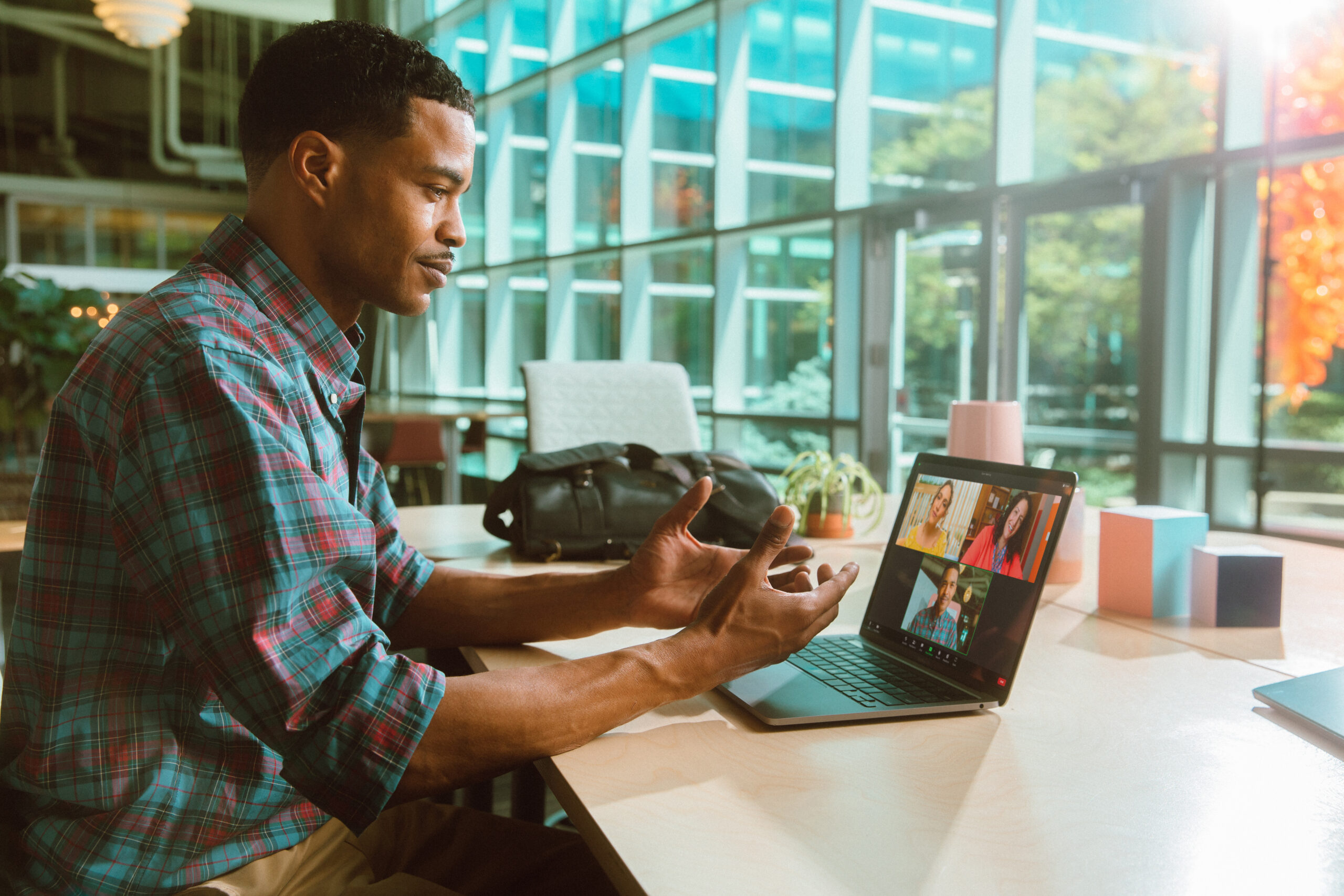 A man in a modern office on a video call, gesturing with hands. Bright windows and colorful decor in the background.