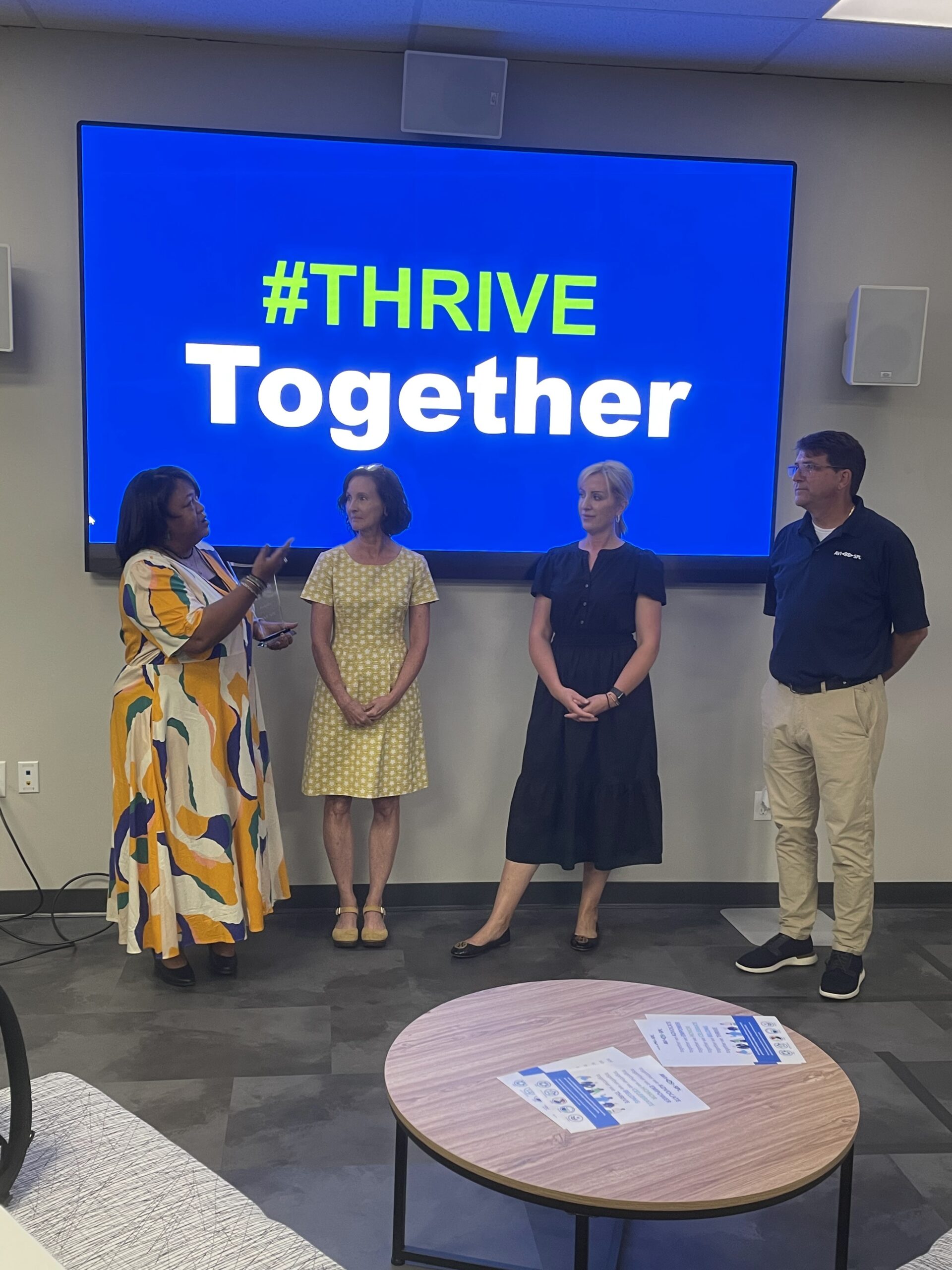 Four people stand before a #THRIVE Together screen; the woman on the left gestures as others listen. A table with papers lies ahead.