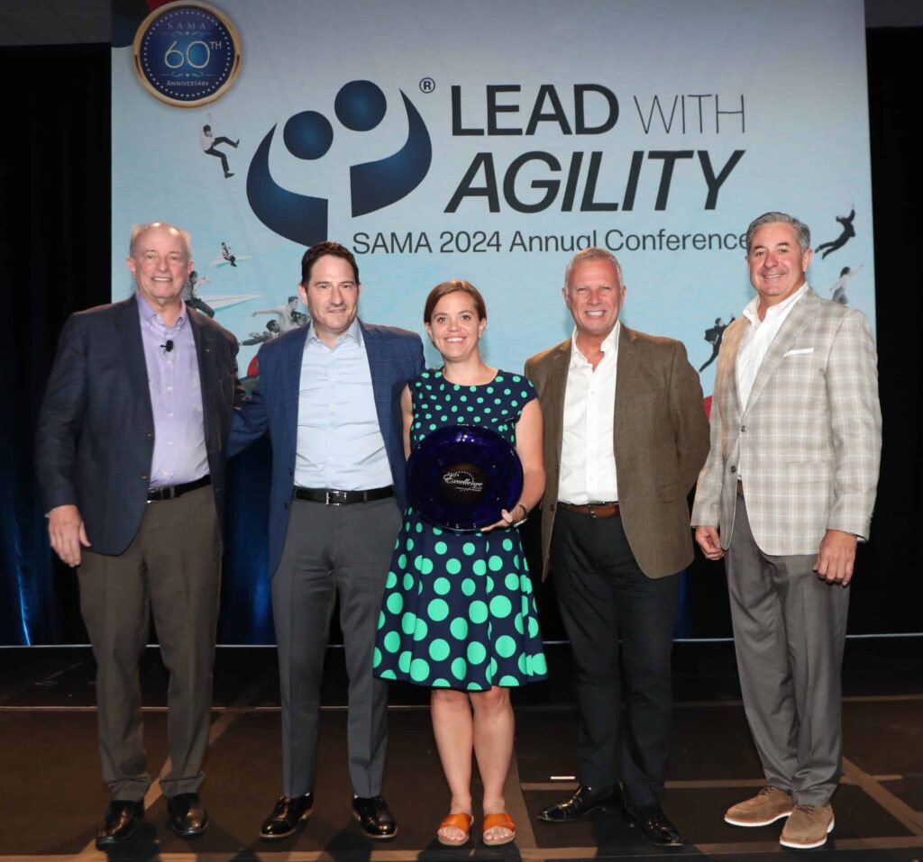 Five people pose at the Lead with Agility SAMA 2024 event; center woman holds an award. All in business attire.