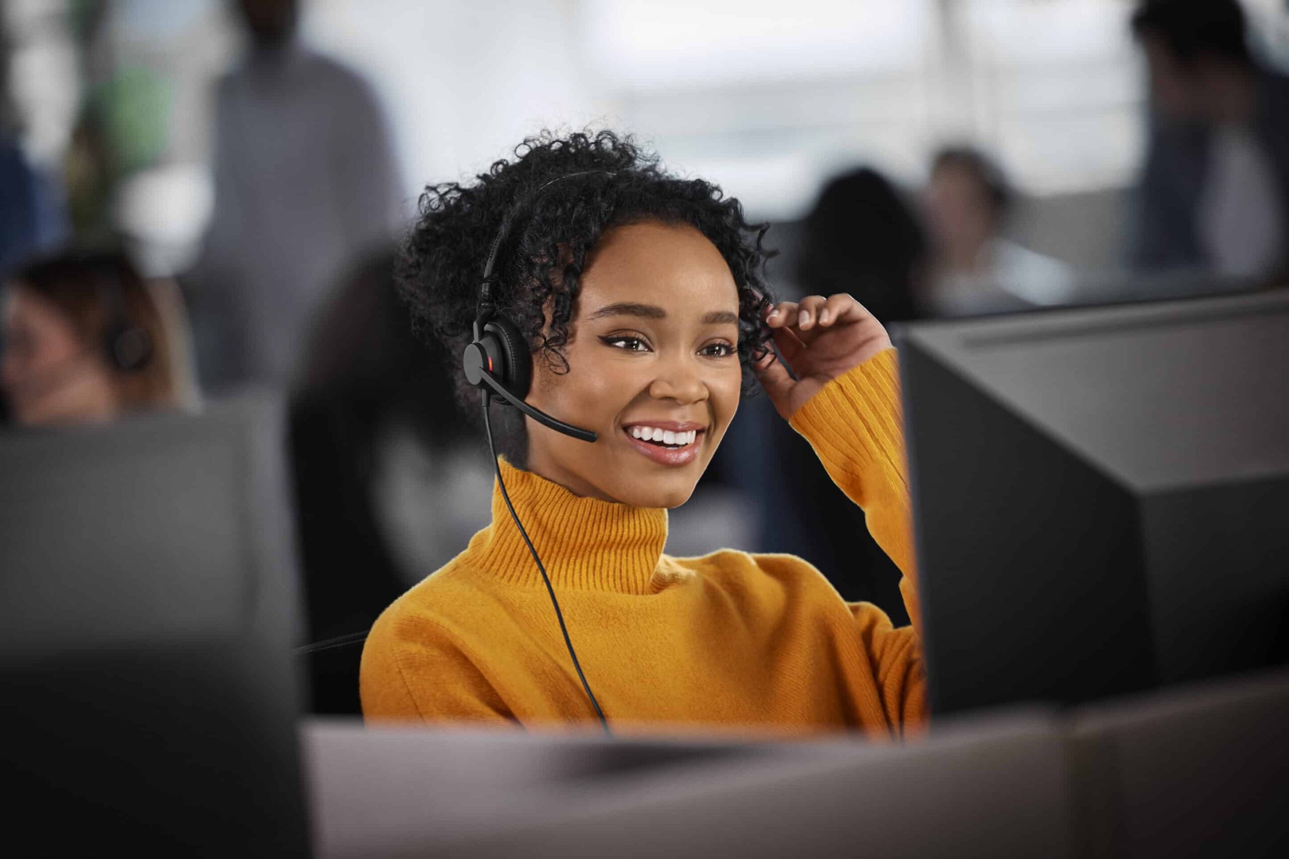 Woman with curly hair in a yellow sweater and headset smiles at a computer in an office, people and desks blurred behind.