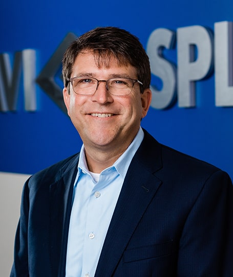 Smiling man in glasses and a navy suit with a light blue shirt stands before a blue wall with partial white lettering.