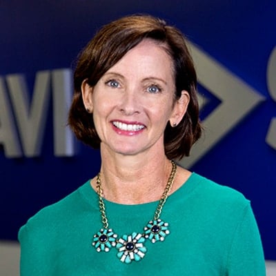 Smiling woman with short brown hair, green top, and beaded necklace against a blue background with partial white text.