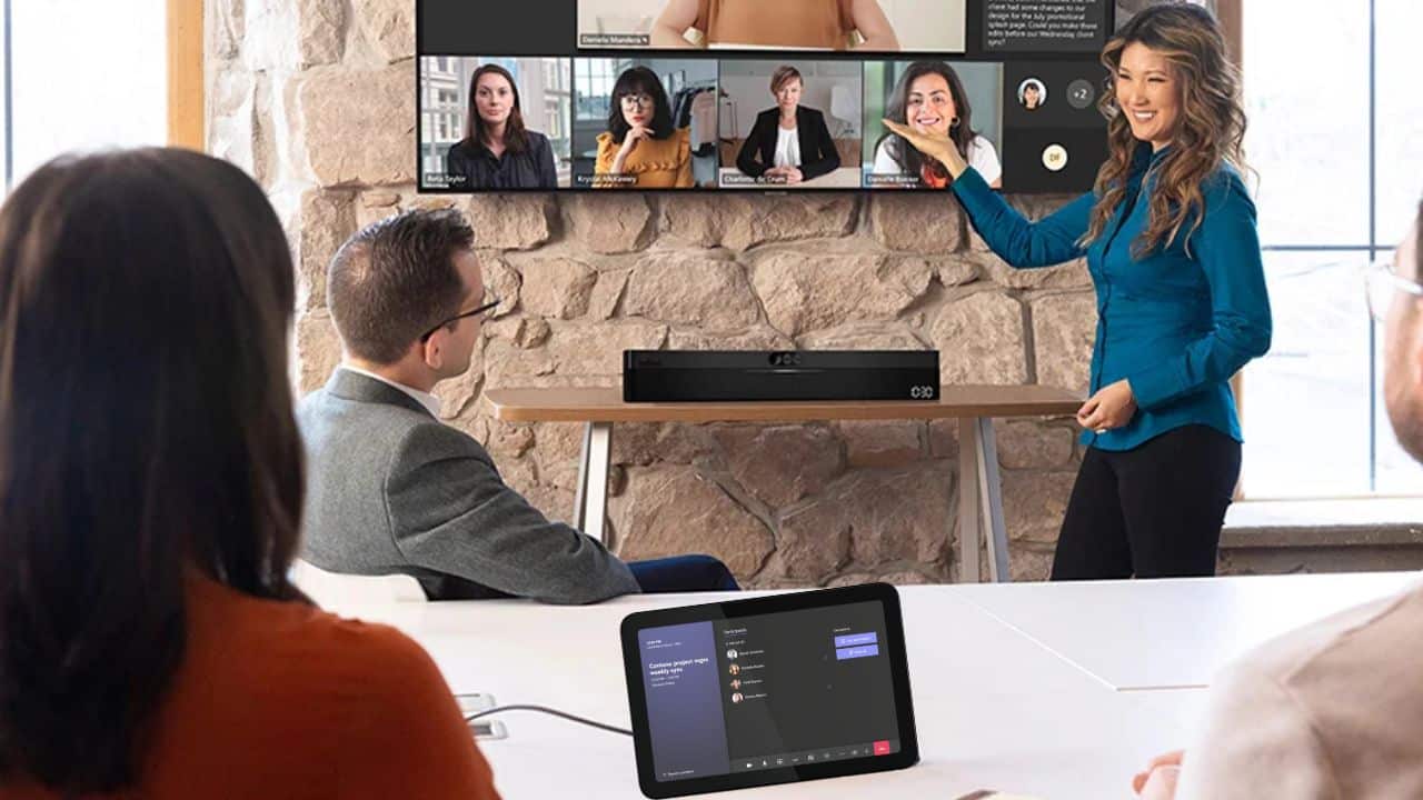 A woman in a blue shirt presents in a conference room, gesturing to a screen with a video call; people watch, tablet on the table.