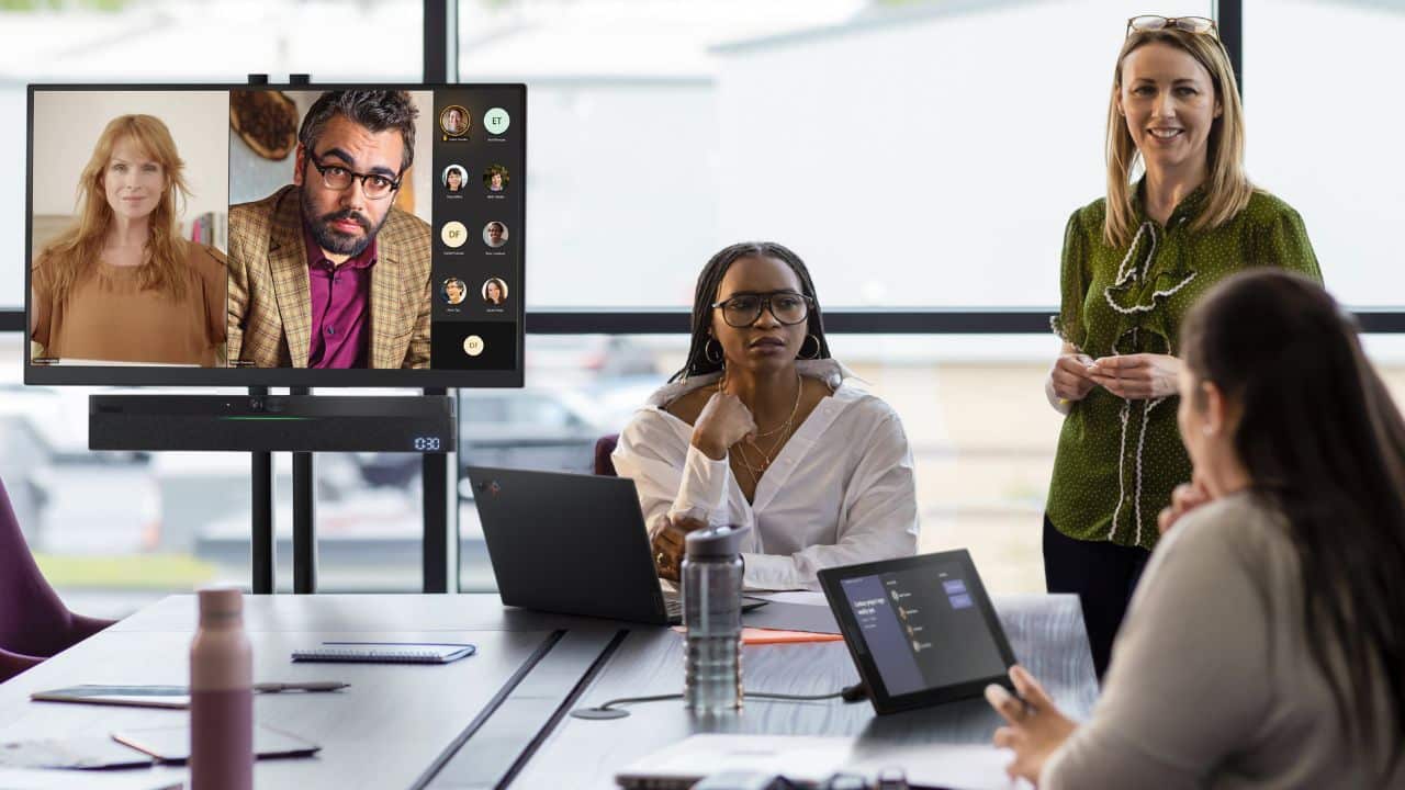 Four people in a meeting; one stands. Two on video call. Laptops, bottles on table. Natural light through large windows.