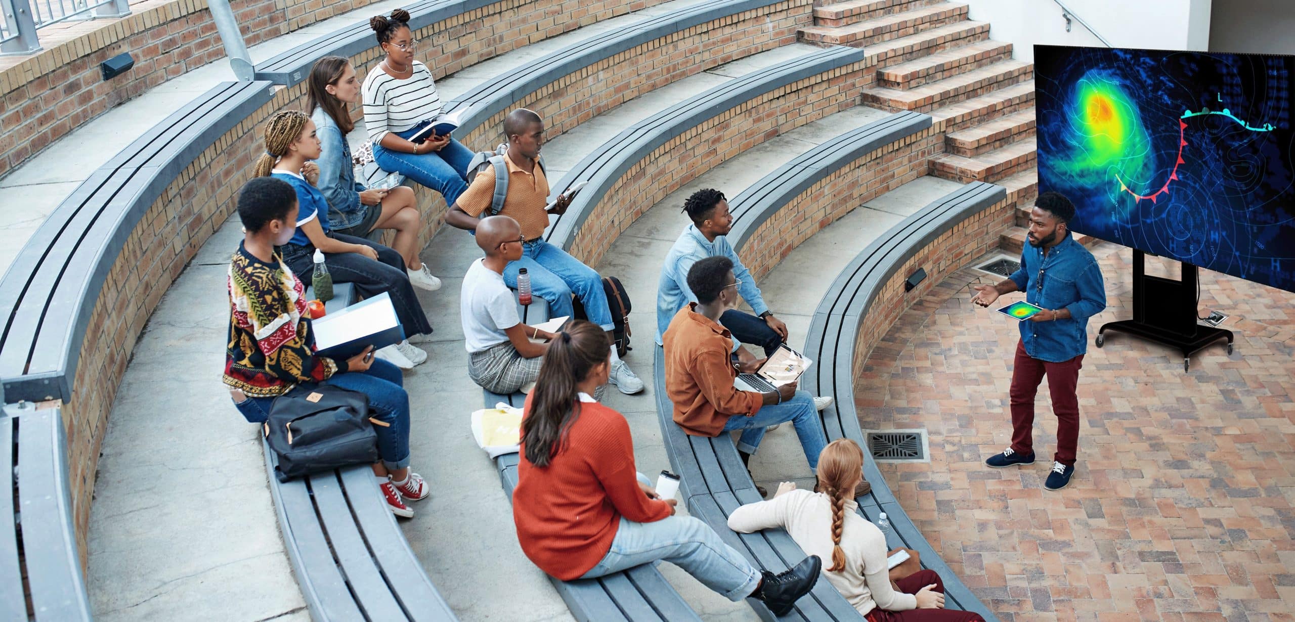 Diverse students sit on amphitheater steps, focused on a presenter by a screen with colorful data graphics.