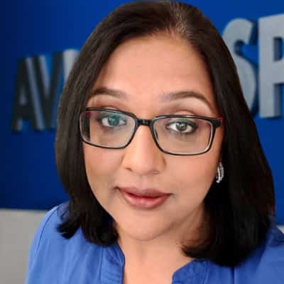 A woman with shoulder-length black hair and glasses, in a blue top, is indoors with a partially visible blue and white background.