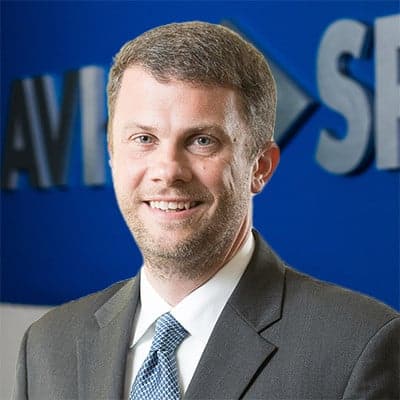 Smiling man in a suit and tie stands before a blue wall, partially obscuring a logo.