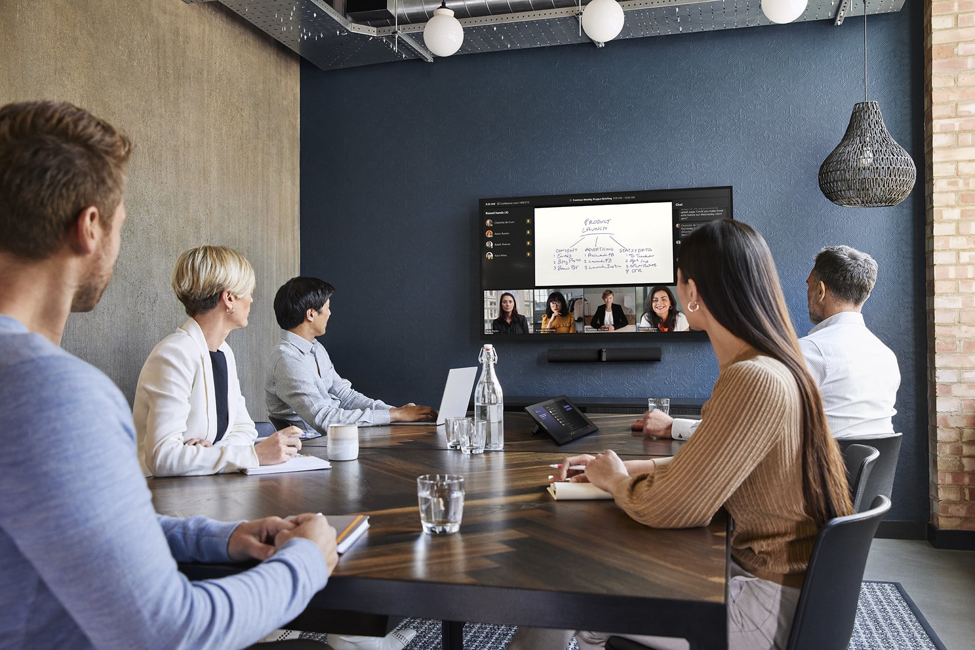 People in a conference room watch a wall screen showing a video call and flowchart; theyre seated with notebooks, laptops, and water.