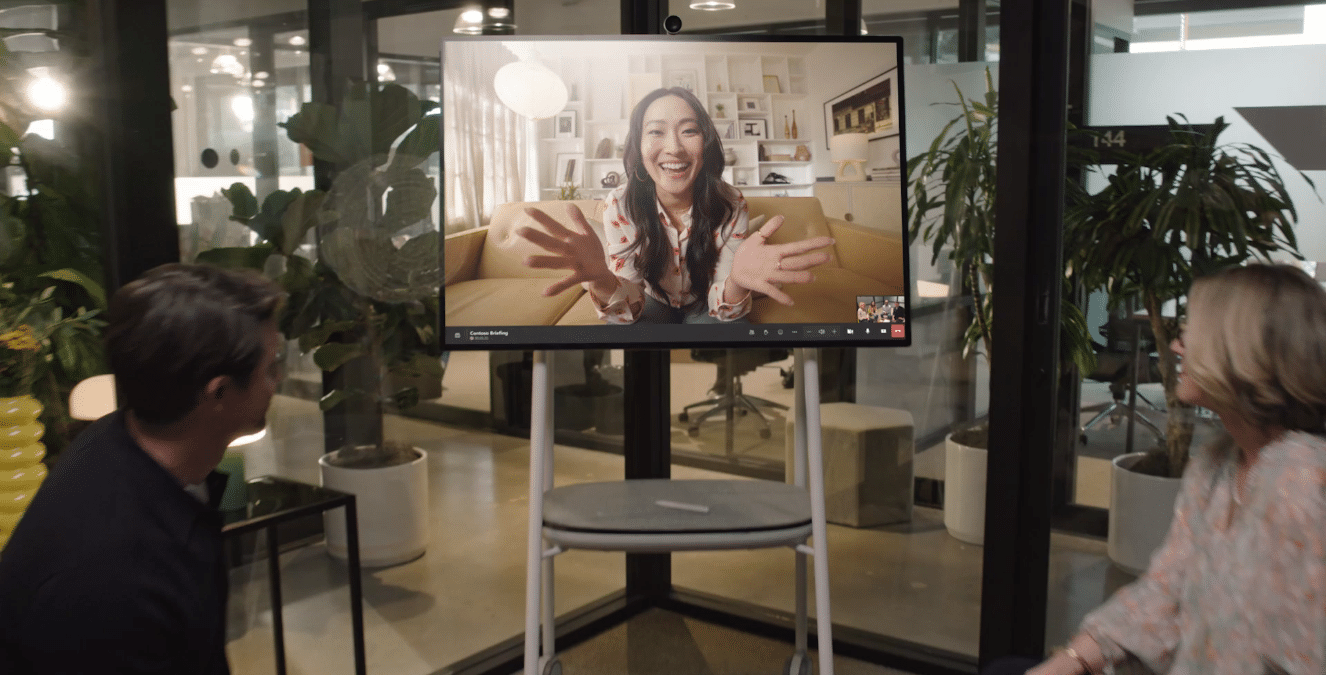 A woman on a screen smiles and gestures energetically during a video call; two nearby people interact enthusiastically with her.