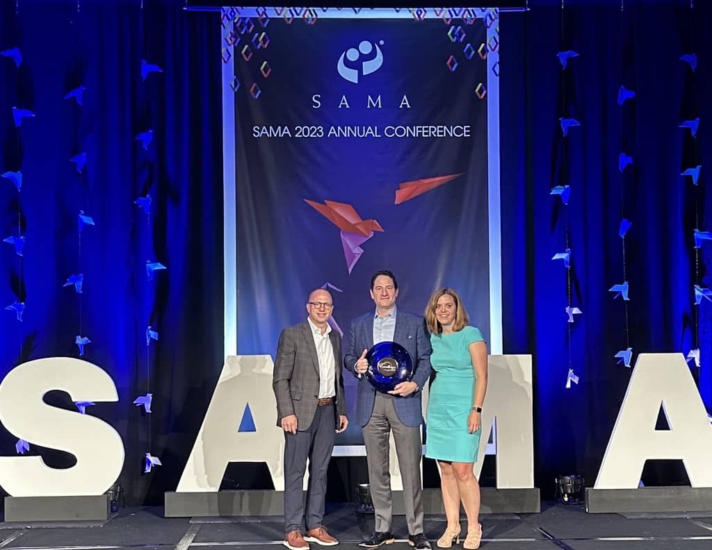 Three people stand on stage at SAMA 2023 Annual Conference, the middle holding a blue award, white SAMA letters in front.