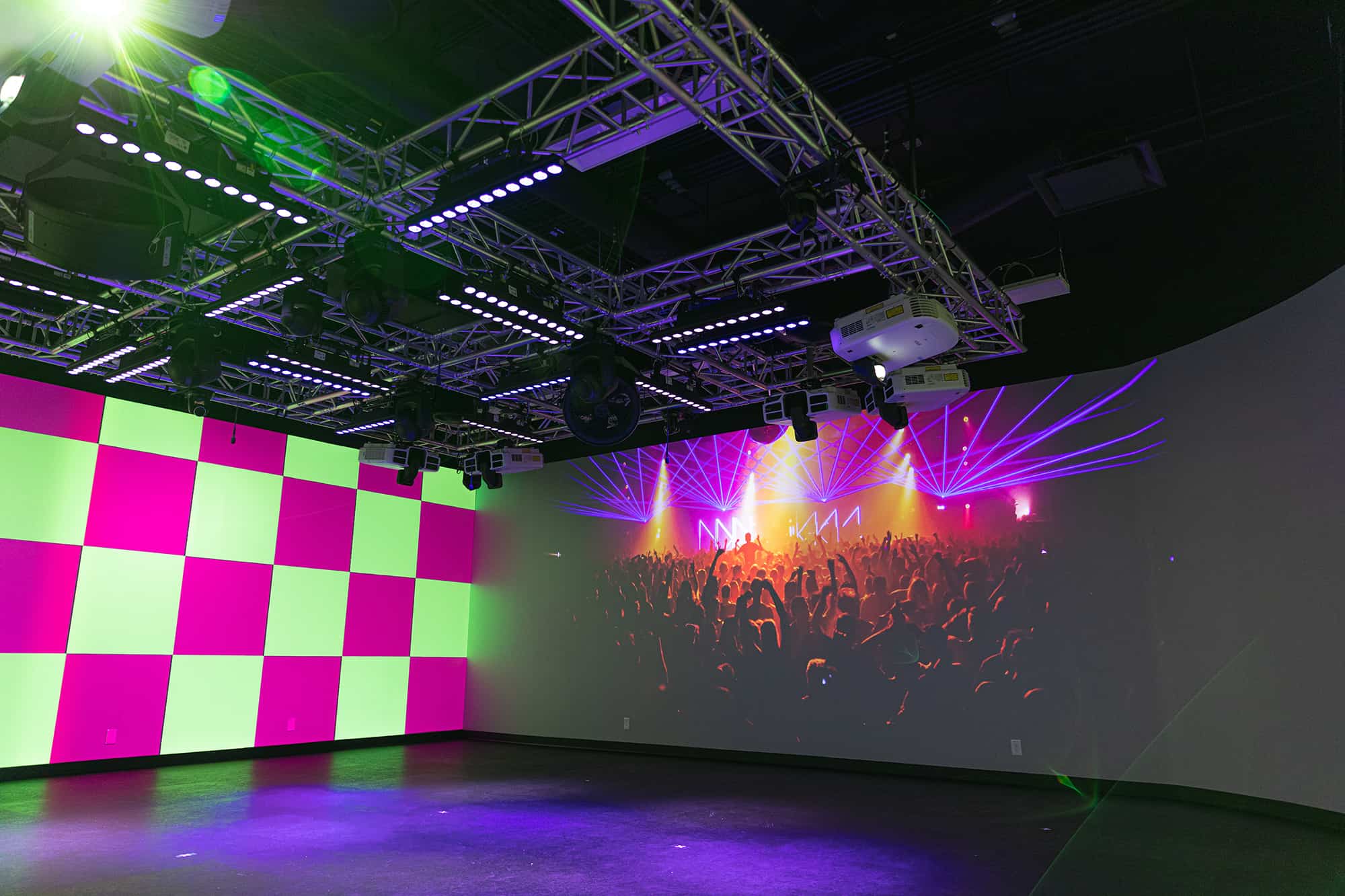 A room with a multi-colored checkerboard wall left, concert scene on large screen right, metal truss ceiling with lights.