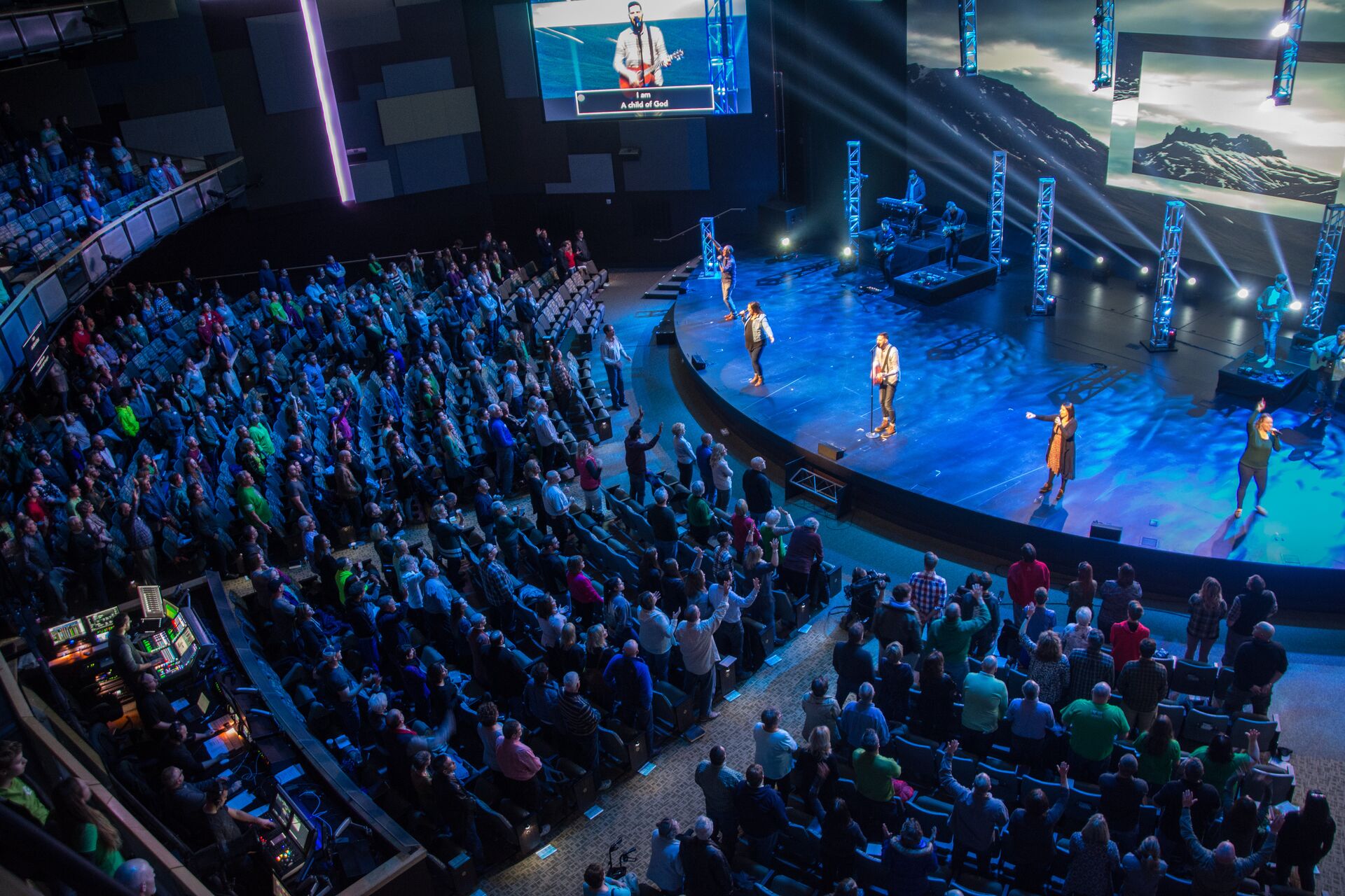 A large audience watches a stage performance with blue lights and mountain backdrop in a modern, spacious auditorium.