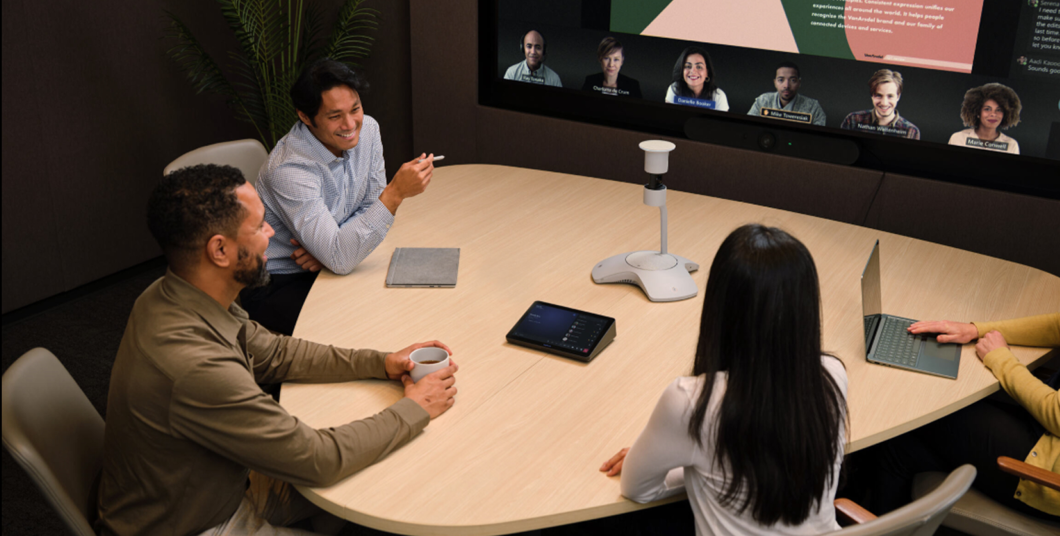 Four people at a conference table with laptops, on a video call displayed on screen; plant and office equipment in background.