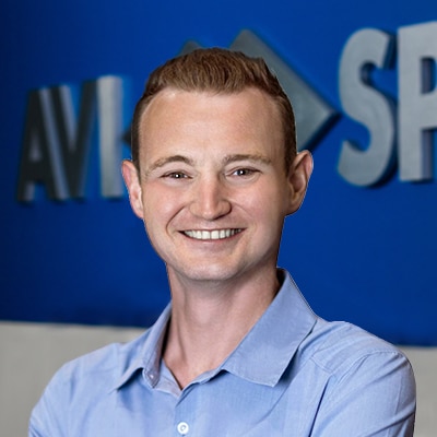 Smiling person with short hair in light blue shirt, stands before a blue wall with blurred text.