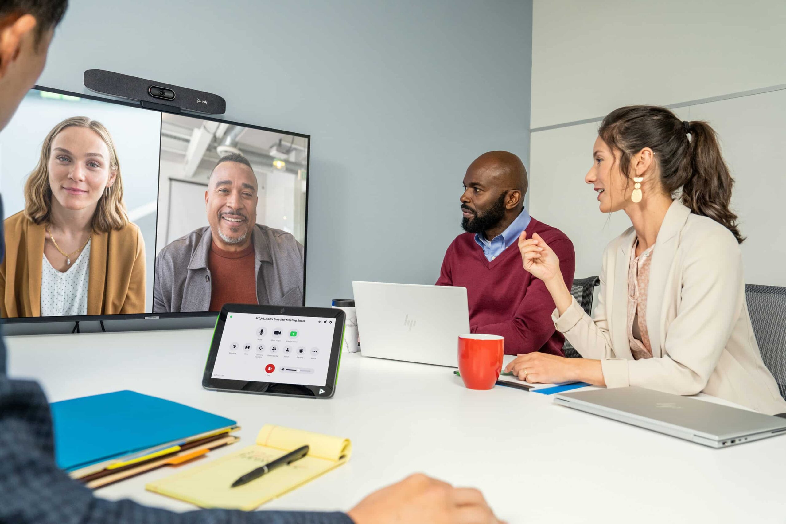 Four people in a meeting room on a video call with two others; laptop, red mug, tablet, and notepad on the table.