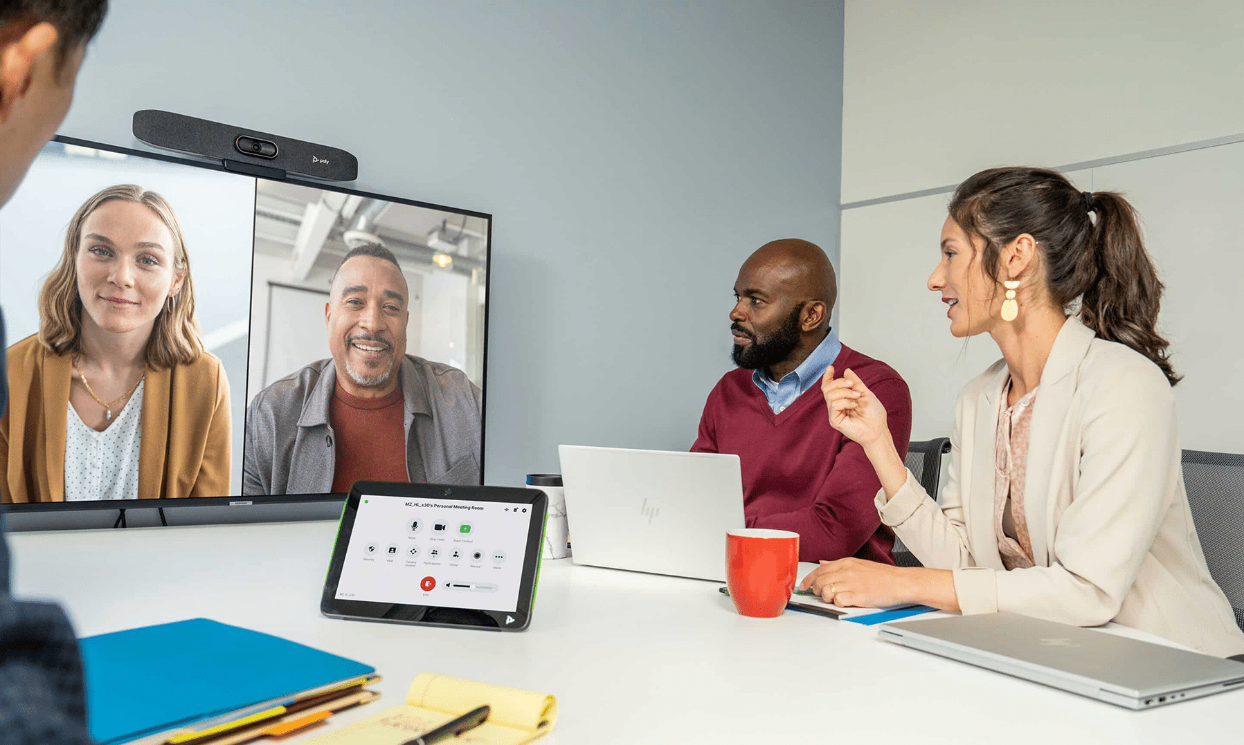 Two on-screen in yellow blazers; two in-room, one with a laptop, the other with a tablet and red cup, actively discussing.