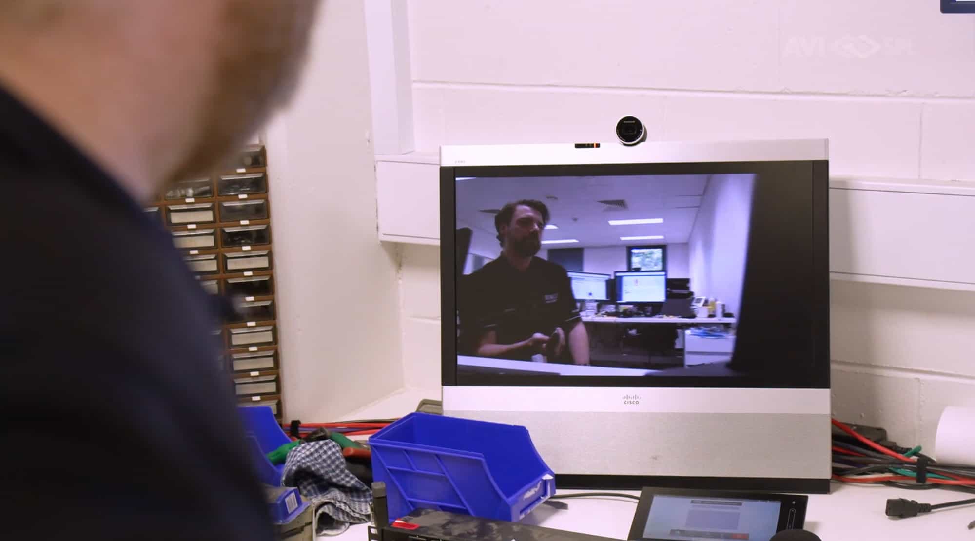 An office worker views a video call showing a person at a desk with dual monitors and visible office supplies.