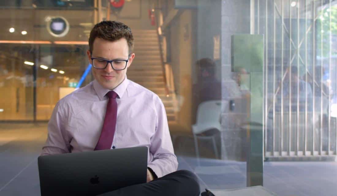 A person in a purple shirt and red tie sits on a bench using a laptop in a modern indoor space with glass walls and stairs.