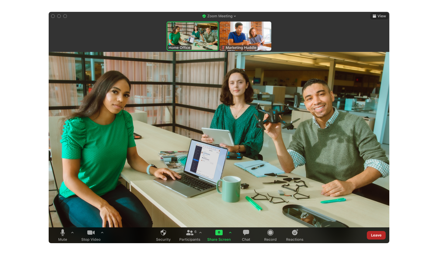 Group video call: diverse trio smiles at camera; laptop, phone, coffee, and office supplies scattered on the table, call icons visible.