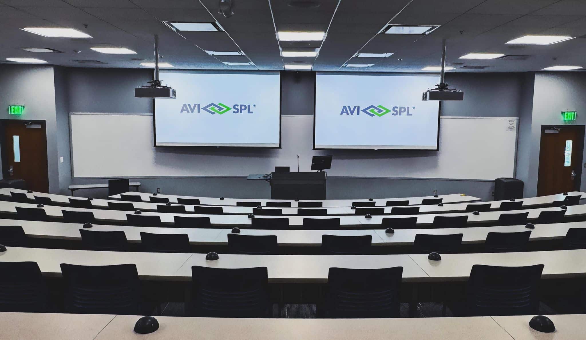 Lecture hall with rows of chairs, tables facing screens showing AVI-SPL logo; overhead lights, exit signs visible.