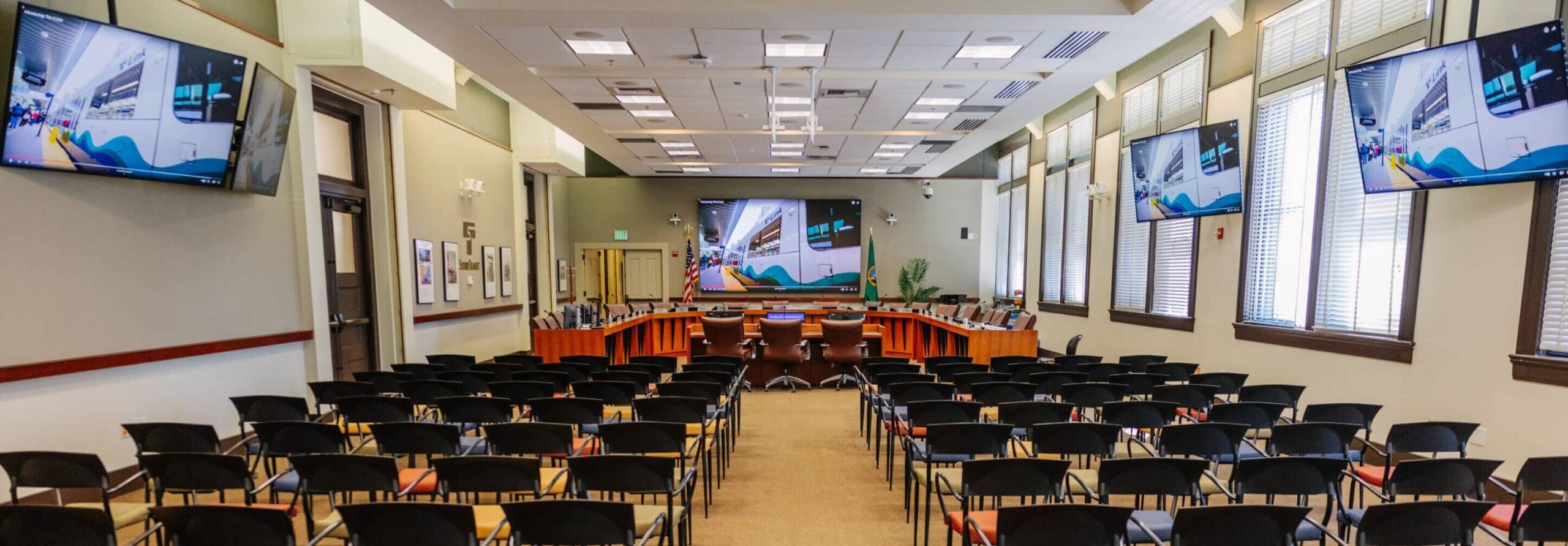 Modern conference room with rows of empty black chairs, large screen, wall-mounted displays, and U-shaped table by tall windows.