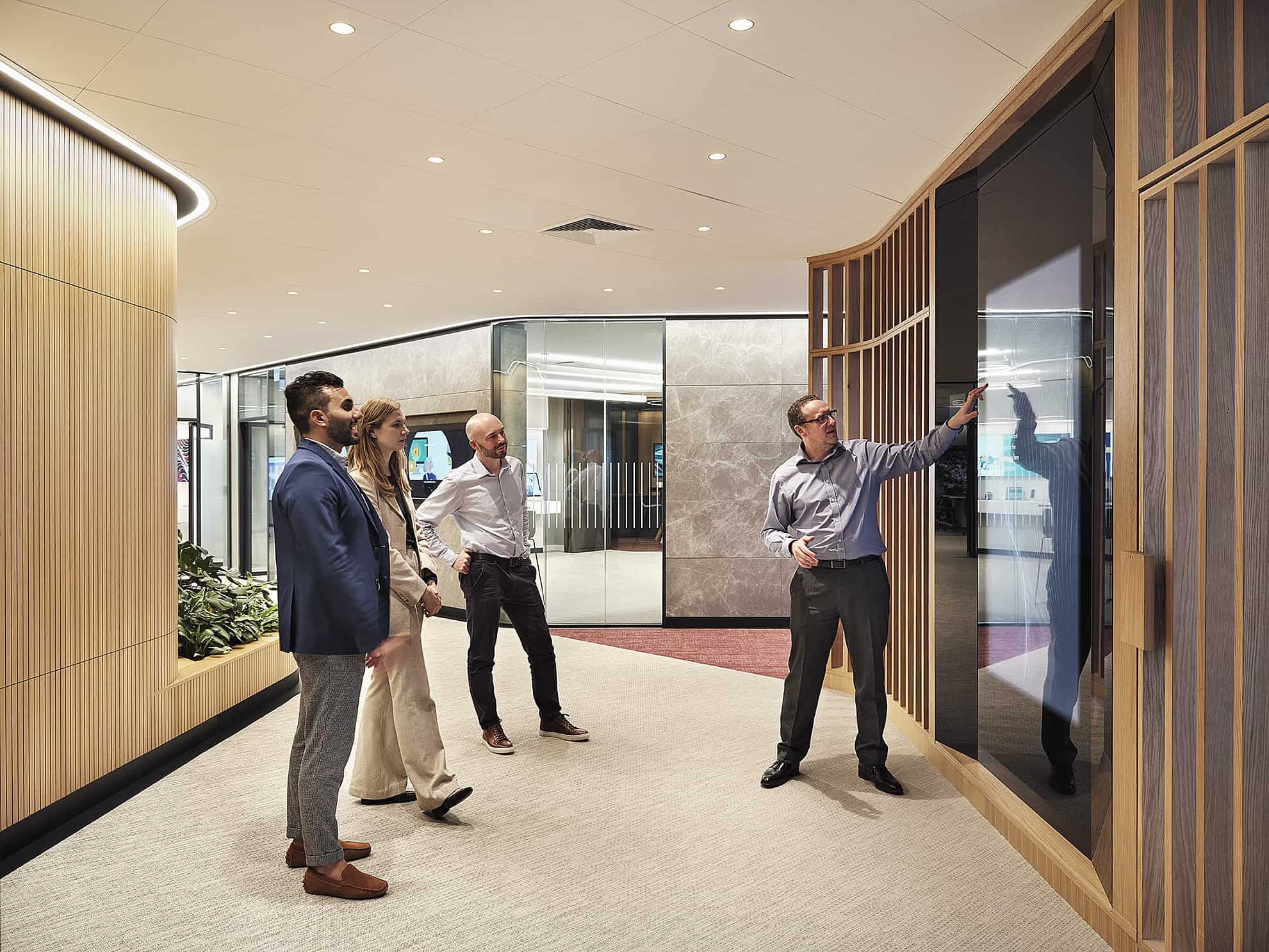 A group of four in a modern office; one points to a screen amid wooden panels, greenery, and glass, fostering collaboration.