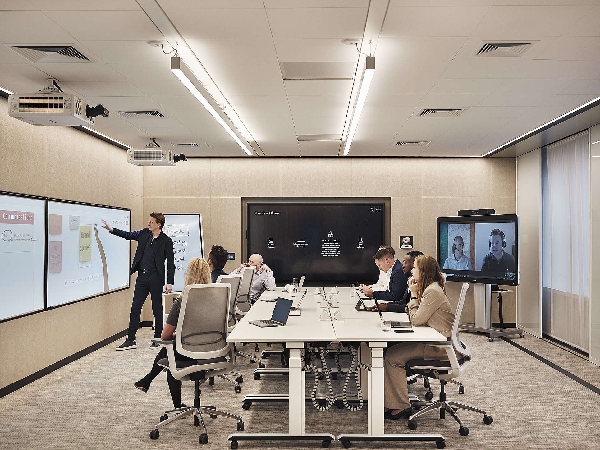 Group at conference table with laptops; one stands by screens, two on video call. Modern, well-lit meeting room.