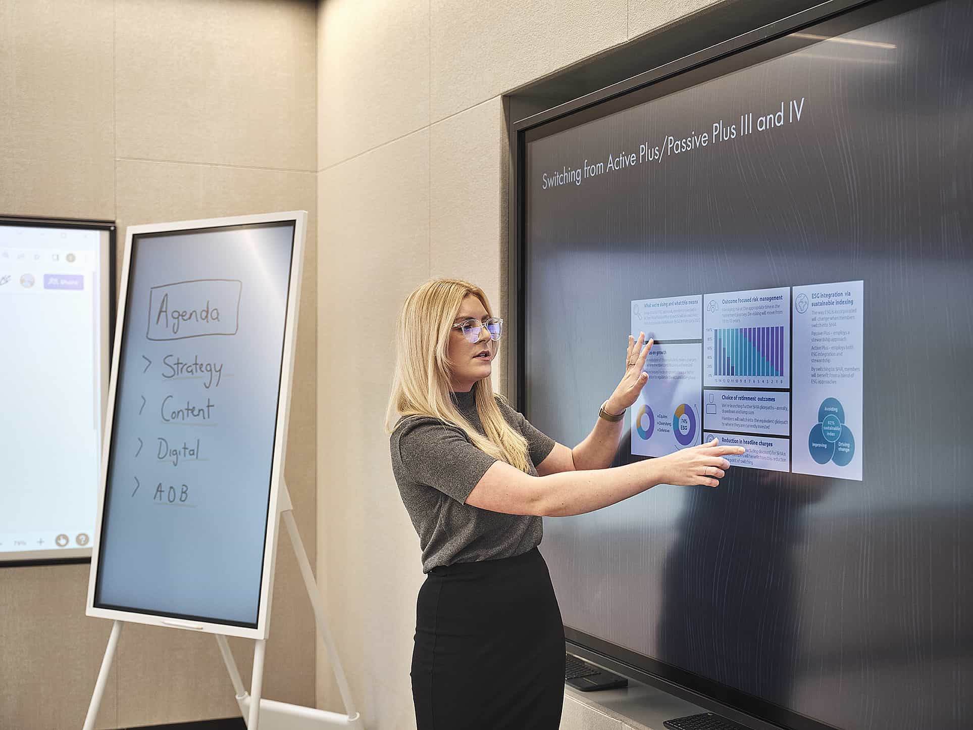 Woman presents in office, pointing at graphs on digital screen; whiteboard with Agenda nearby.