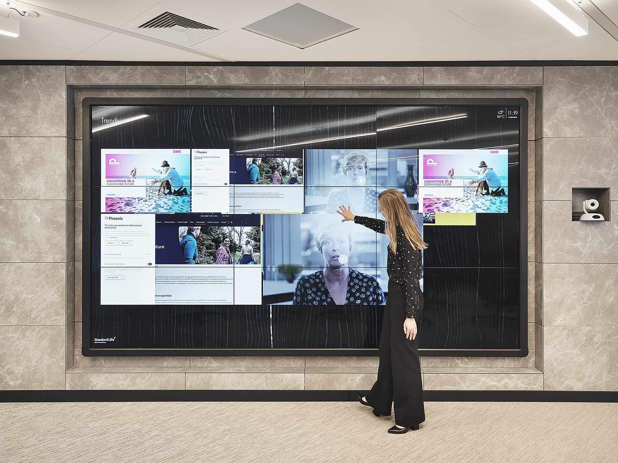 A woman in black interacts with a large touchscreen display in an office, touching images and documents.